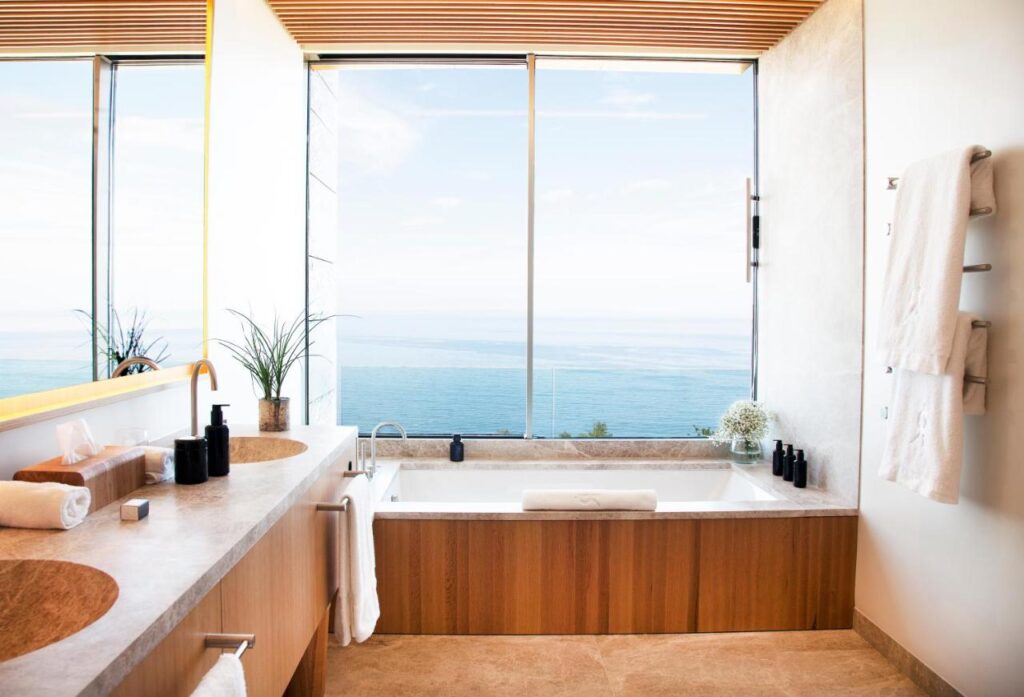 Best Boutique Hotels in San Sebastian 25 Modern bathroom inspired by boutique hotels in San Sebastian, with large windows overlooking the ocean, a wooden bathtub, double sink countertop, rolled towels, black toiletries, green plants, and white towels hanging on the wall.