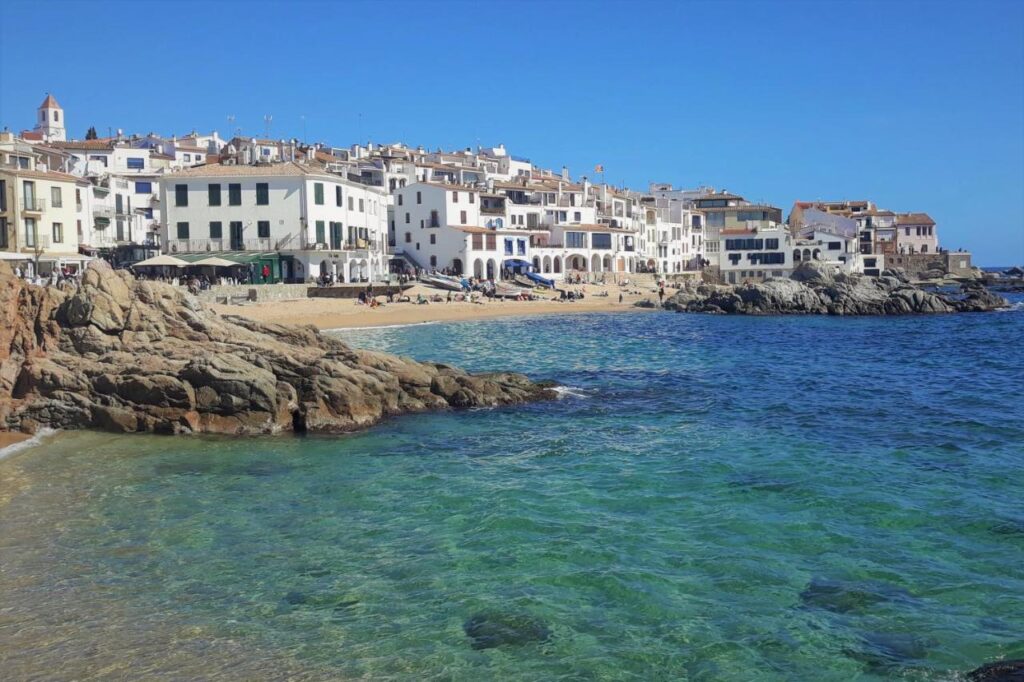 A coastal village with white buildings and tiled roofs lines a sandy beach. Clear blue-green water meets rocky shorelines under a bright sky. People relax on the beach, waterfront cafes, and chic boutique hotels in Costa Brava.