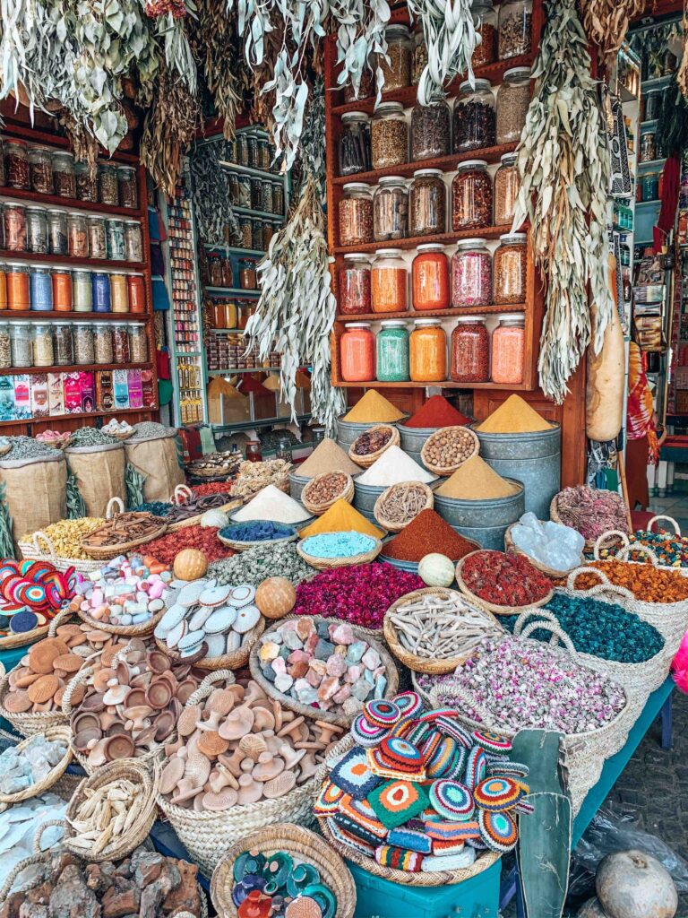 12 Charming Budget Riads in Marrakech 2 A vibrant market stall displays piles of colorful spices, dried herbs, and woven baskets filled with goods—a scene often found near budget riads in Marrakech. Jars line the wooden shelves, and dried plants hang above, enriching the lively and aromatic atmosphere.