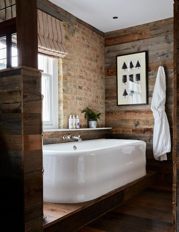 15 Best Boutique Hotels in London, UK 36 A rustic bathroom with exposed brick and wood walls, a white bathtub, a striped Roman shade, and boutique hotels in London-inspired touches like a crisp white robe and elegant toiletries beside a potted plant.