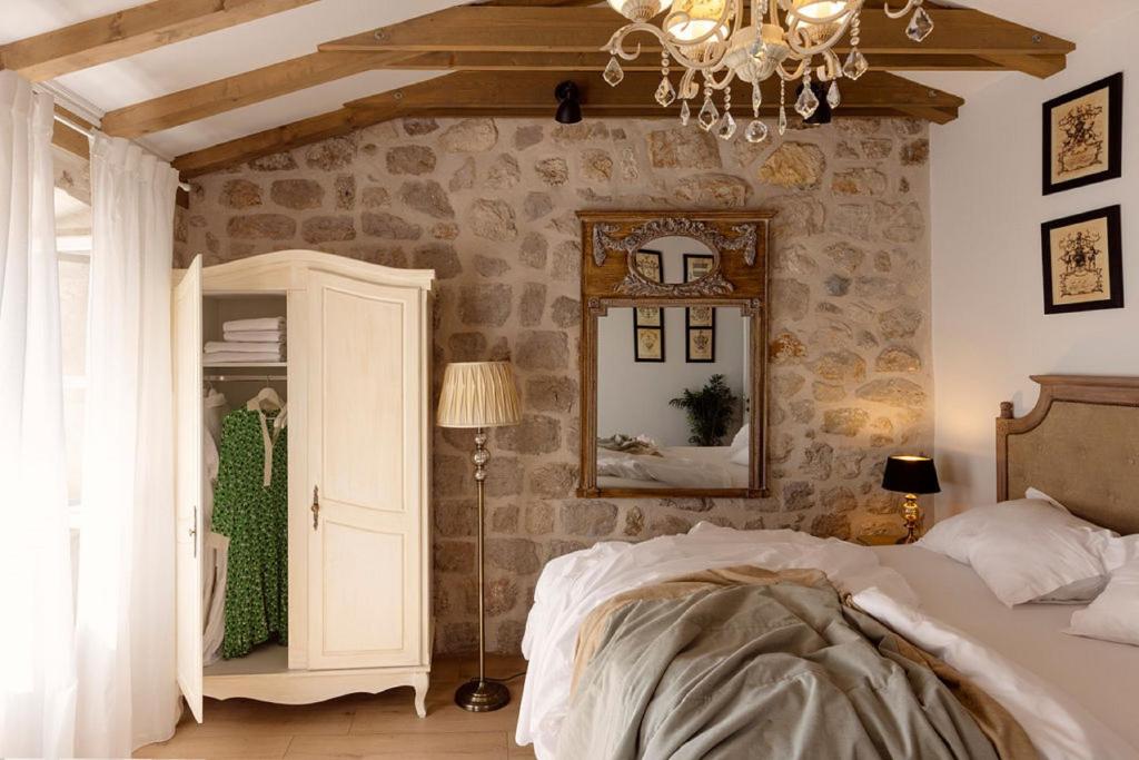 Most Unique Boutique Hotels in Dubrovnik 26 A cozy bedroom in the style of Boutique Hotels in Dubrovnik, with stone walls, a wooden beam ceiling, a double bed with white and beige bedding, an open wardrobe showing a green dress, an ornate mirror, and soft chandelier lighting.