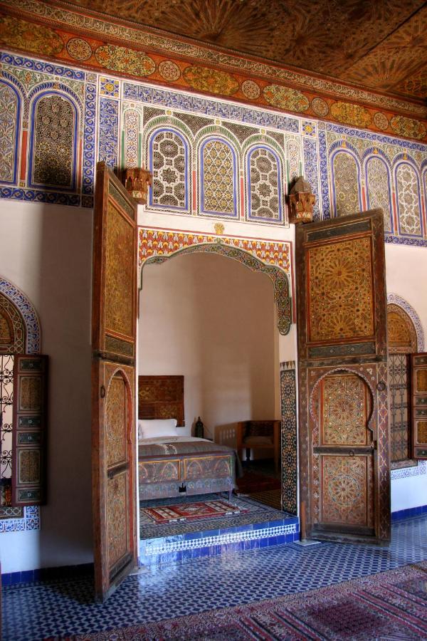 7 UNIQUE Riads in Fez You Won't Forget 3 Ornate Moroccan interior with colorful geometric tilework, intricate wooden doors, and arched doorways typical of riads in Fez. Detailed wall patterns, a bed, and rugs are visible through the open doors into the adjacent room.