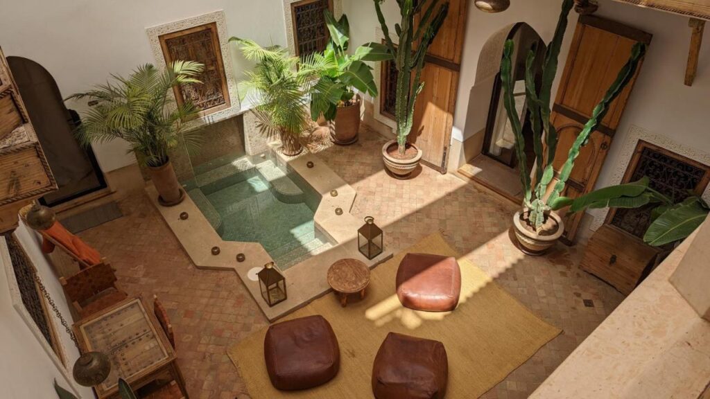 12 Charming Budget Riads in Marrakech 43 Sunlit courtyard at one of the budget riads in Marrakech, featuring a central pool, potted tropical plants, lanterns, three brown ottomans on a woven rug, and wooden furniture amid tiled floors and detailed windows.