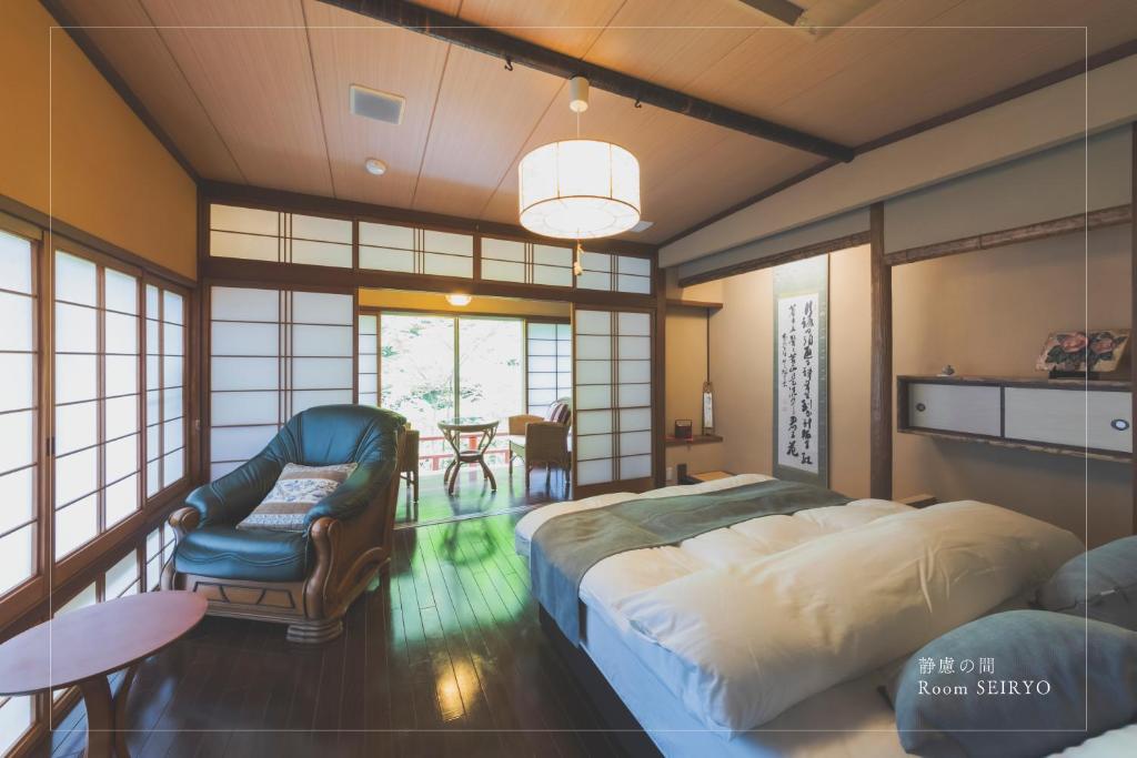 8 BEST Ryokans in Osaka 32 A spacious traditional Japanese-style room reminiscent of ryokans in Osaka, featuring twin beds, a cushioned armchair, shoji sliding doors, a calligraphy scroll, and a seating area by large windows overlooking lush greenery.