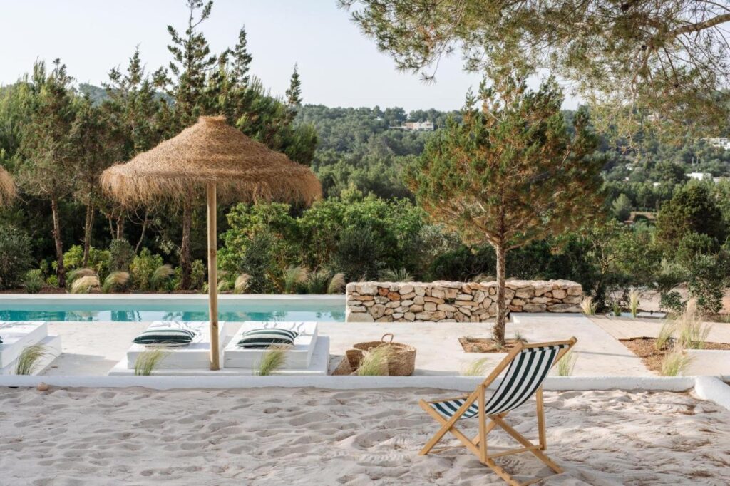 10 AUTHENTIC Finca Hotels in Ibiza 2 A sandy area with a striped deck chair faces a pool lined with lounge chairs and straw umbrellas, surrounded by trees and greenery, evoking the peaceful charm of finca hotels in Ibiza, with a stone wall and distant hills in the background.
