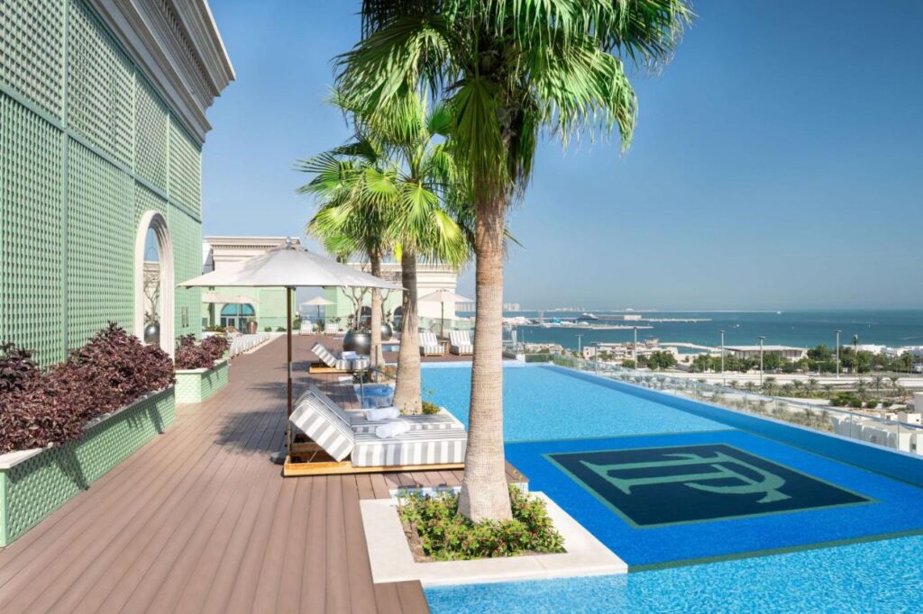 Where to Stay in Doha, Qatar: Transit, City & Beach Hotels 28 A rooftop swimming pool with a large logo, lined with palm trees and sun loungers, overlooks a cityscape and the sea under a clear blue sky—the perfect spot when searching for where to stay in Doha.