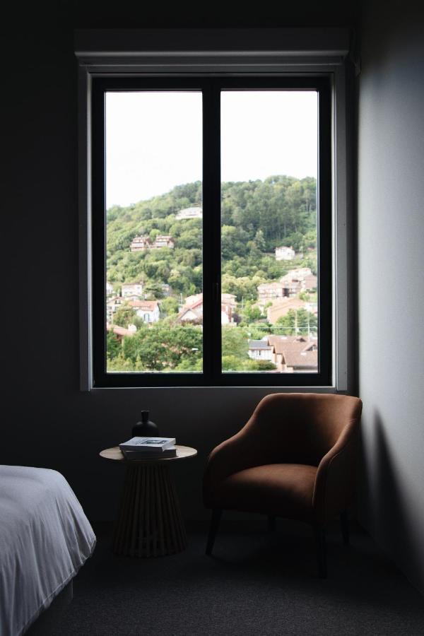 Best Boutique Hotels in San Sebastian 30 A large window in a dimly lit room frames a scenic view of green hills and houses, reminiscent of boutique hotels in San Sebastian. Inside, a brown armchair, round table with a book, and part of a white bed create an inviting retreat.