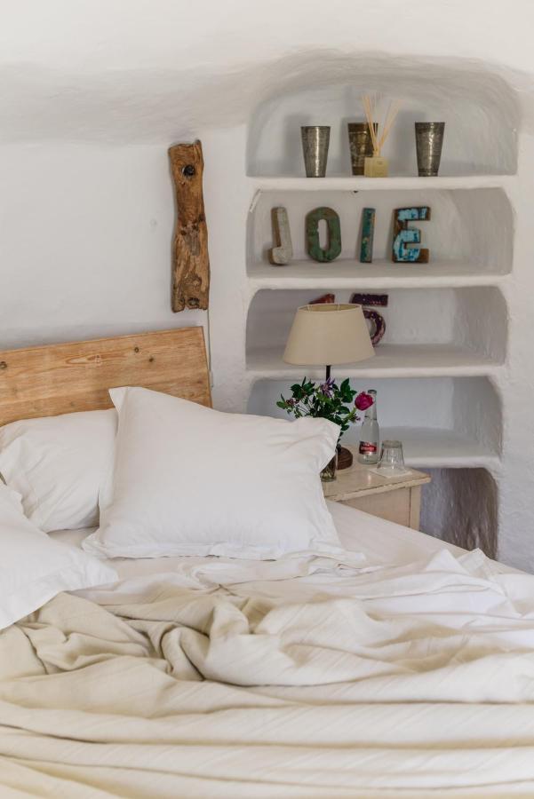 10 AUTHENTIC Finca Hotels in Ibiza 22 A cozy bed with white pillows and beige sheets sits beside a nightstand with a lamp and flowers, evoking the relaxed charm of finca hotels in Ibiza. Built-in wall shelves display metal cups and colorful letters spelling JOIE.