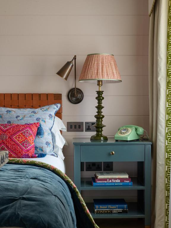 15 Best Boutique Hotels in London, UK 49 A cozy bedroom scene inspired by boutique hotels in London, featuring a bed with colorful patterned pillows and a blue blanket beside a blue nightstand holding a green lamp, a vintage rotary phone, and several books on the lower shelf.