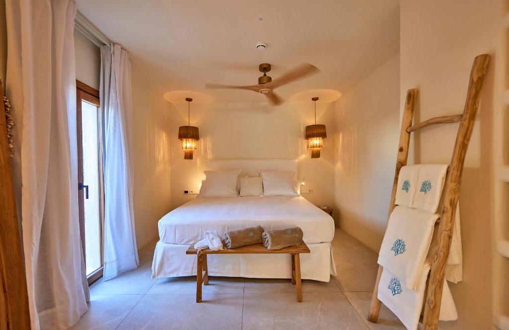 A cozy bedroom inspired by boutique hotels in Formentera, featuring a large bed with white linens, two hanging bedside lamps, a ceiling fan, and a wooden bench. A ladder with towels leans against the wall and natural light pours in through the glass door.
