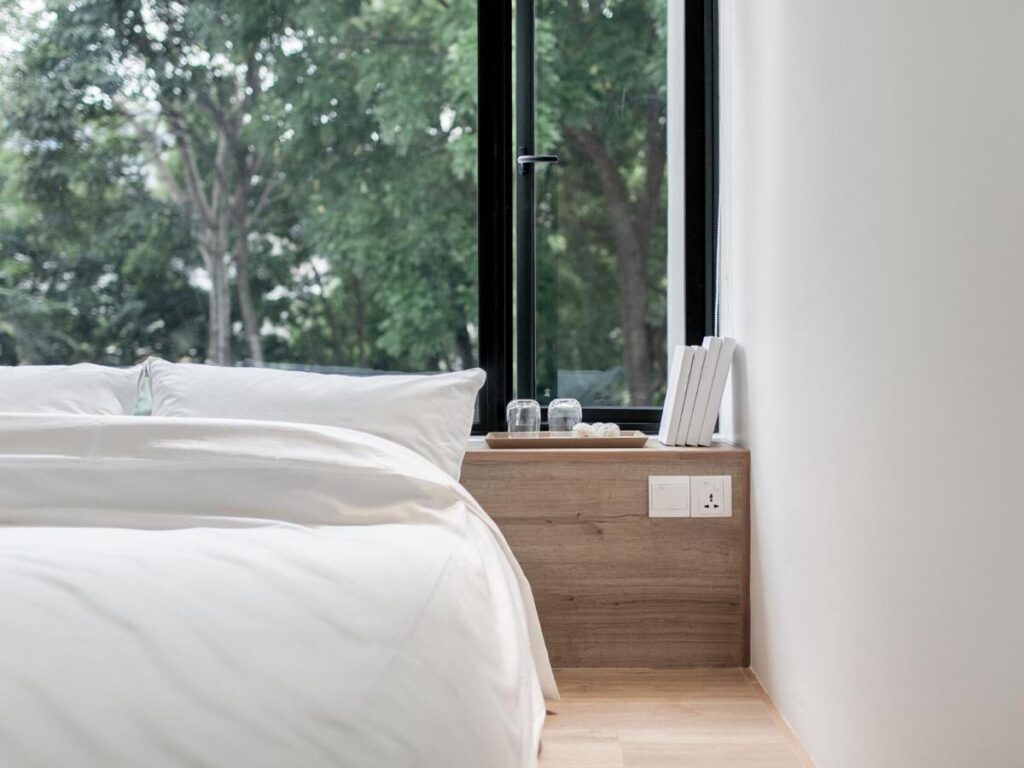 13 Most Unique Hotels in Singapore for an Incredible Holiday 15 A minimal bedroom with a neatly made bed in white linens beside a large window showing green trees—perfect for those seeking unique hotels in Singapore. On a wooden bedside shelf are books, glass cups, and an electrical outlet.