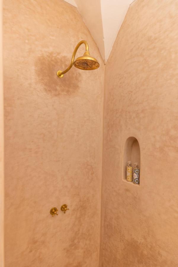 12 Charming Budget Riads in Marrakech 27 A tiled shower reminiscent of budget riads in Marrakech features a gold showerhead and two gold knobs. A recessed shelf in the textured beige wall holds two patterned bottles, evoking a touch of Moroccan charm.