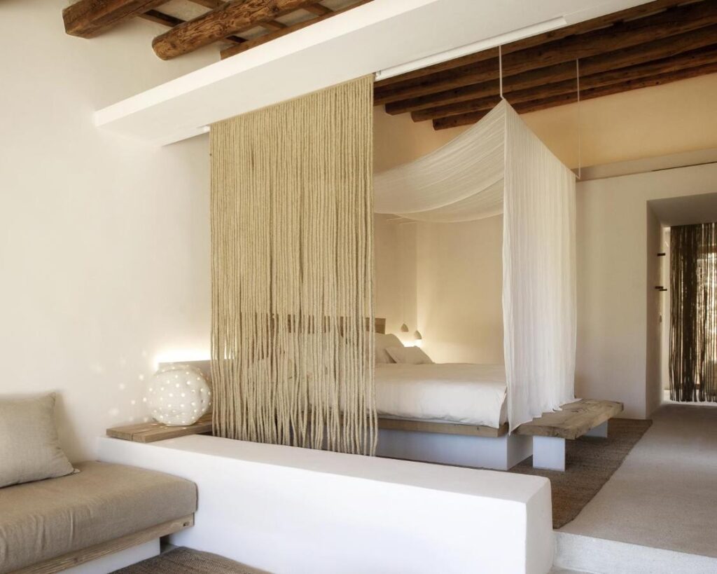Unique Agroturismos in Mallorca 24 A minimalist bedroom inspired by agroturismos in Mallorca features a bed with a white canopy, beige fringe curtain, exposed wooden beams, neutral tones, and a built-in cushioned bench in the foreground.