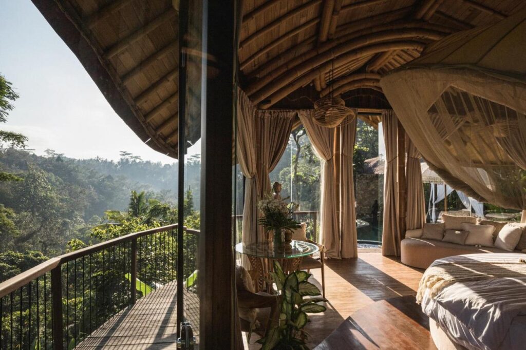 13 Most UNIQUE Places to Stay in Ubud 21 A cozy bedroom with a canopy bed and wooden ceiling opens onto a balcony overlooking lush green trees and a distant forested valley—one of the unique places to stay in Ubud, bathed in soft morning light.