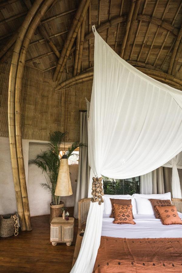 13 Most UNIQUE Places to Stay in Ubud 18 A cozy bedroom with a high bamboo ceiling, canopy bed draped in white fabric, brown patterned bedding, and soft natural light—one of the unique places to stay in Ubud for a relaxing retreat.