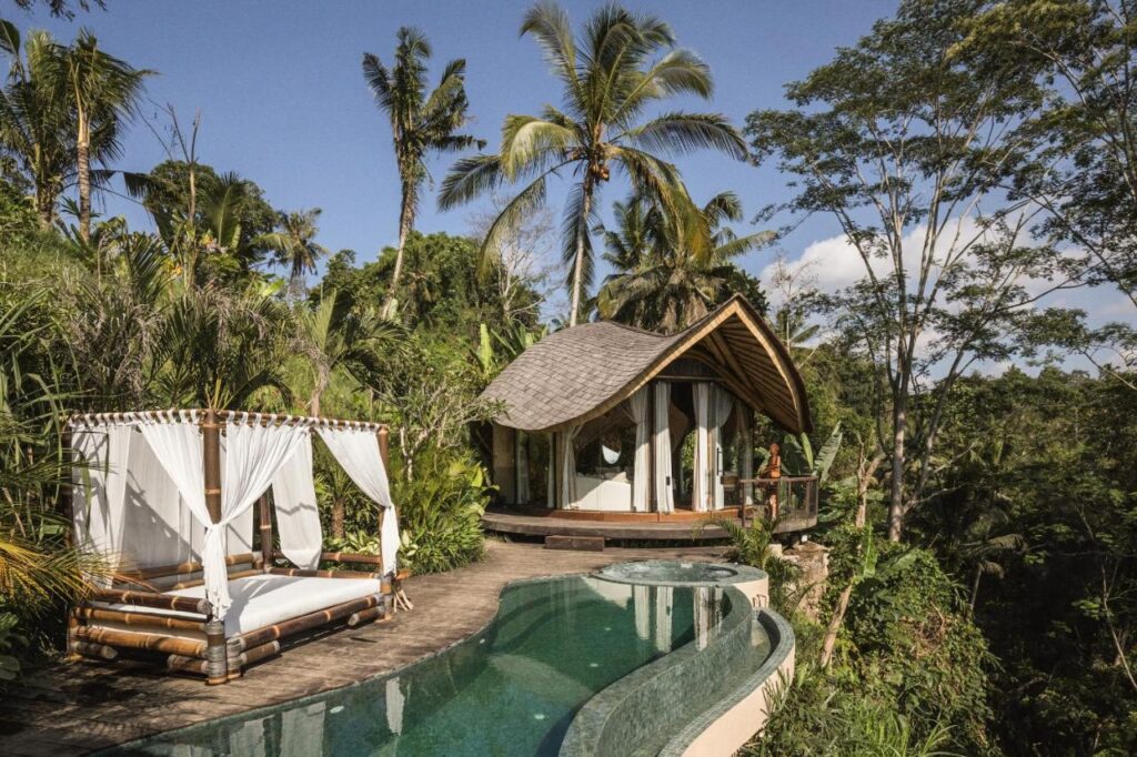 13 Most UNIQUE Places to Stay in Ubud 20 A tropical villa with a curved wooden roof, set amid lush greenery. One of the unique places to stay in Ubud, it features an infinity pool and a bamboo canopy daybed with white curtains, surrounded by palm trees and dense forest under a clear blue sky.