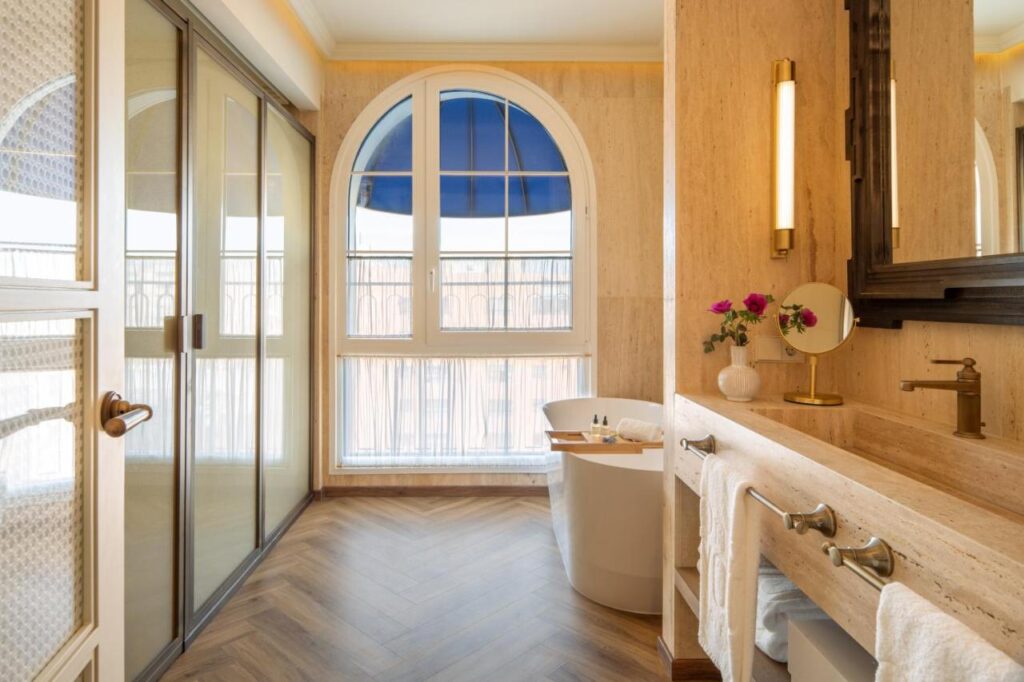 10 UNIQUE Hotels with Pool in Seville 28 Elegant bathroom with a freestanding bathtub by a large arched window, double sink, mirror, flowers in a vase, and a walk-in shower. Warm lighting and wood-patterned floor evoke the luxury found in top hotels with pool in Seville.