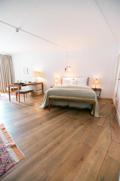 Authentic Boutique Hotels in Munich 12 A cozy, modern bedroom inspired by boutique hotels in Munich, featuring a large bed, wooden floors, a desk and chair near a window with beige curtains, and soft lighting from bedside lamps.