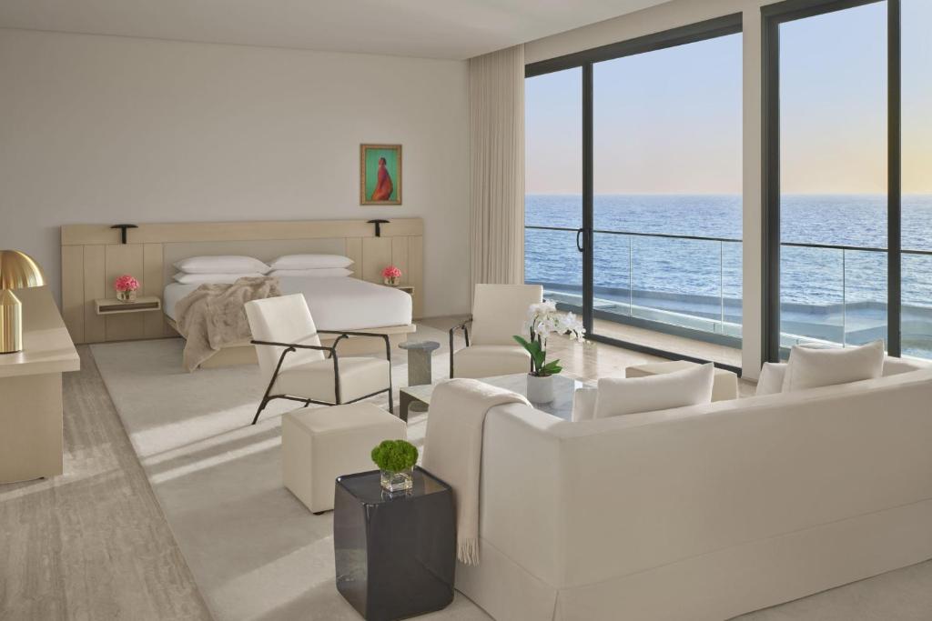 9 Best Hotels in Saudi Arabia: Unique Stays Across the Country 9 A modern, spacious hotel room with cream-colored furniture, a bed, and seating area. Large floor-to-ceiling windows overlook a balcony and ocean view at sunset. Among the Best Hotels in Saudi Arabia, it features a colorful painting above the bed.