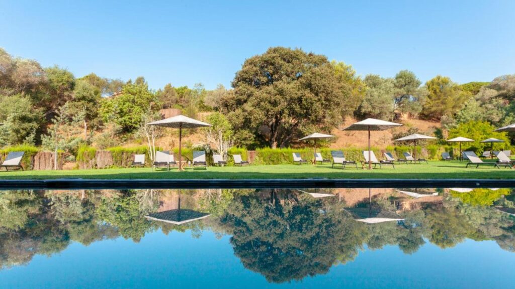 A row of lounge chairs and white umbrellas sit on a grassy lawn beside a still swimming pool at one of the boutique hotels in Costa Brava, with trees and hills reflected in the water under a clear blue sky.