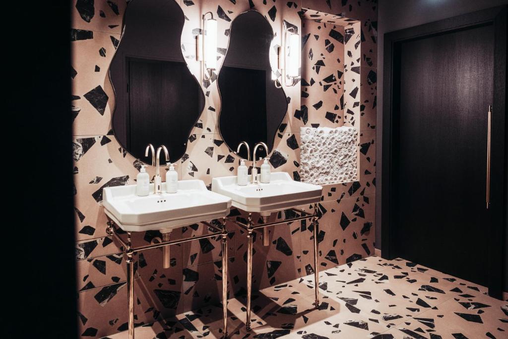 Unique Boutique Hotels in Salzburg 22 A modern bathroom inspired by boutique hotels in Salzburg, with terrazzo-patterned walls and floor, two white sinks on metallic legs, irregular mirrors, wall-mounted lights, and a dark wooden door.
