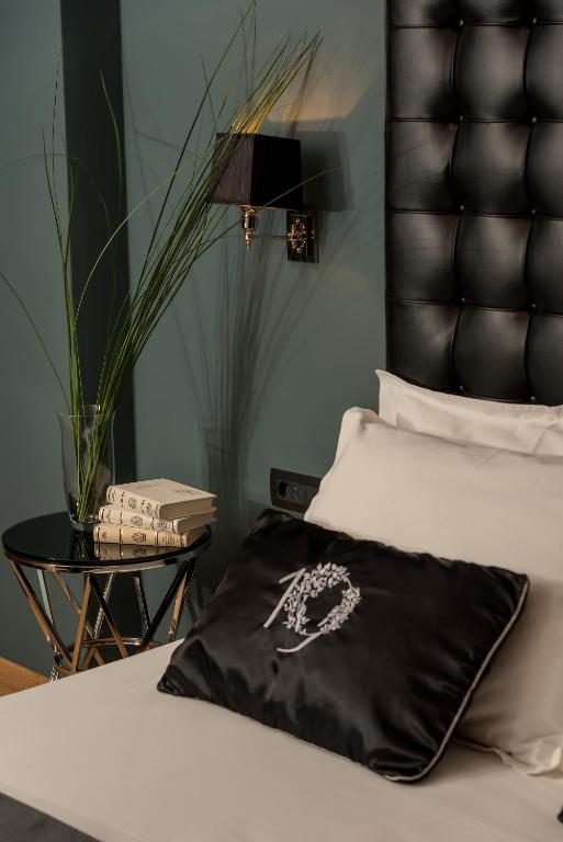 8 TOP Boutique Hotels in Split 35 A stylish bedroom, reminiscent of boutique hotels in Split, features a black tufted headboard, white bedding, a decorative pillow with a wreath design, a modern glass nightstand with books and tall grass in a vase, and a wall-mounted black lamp.