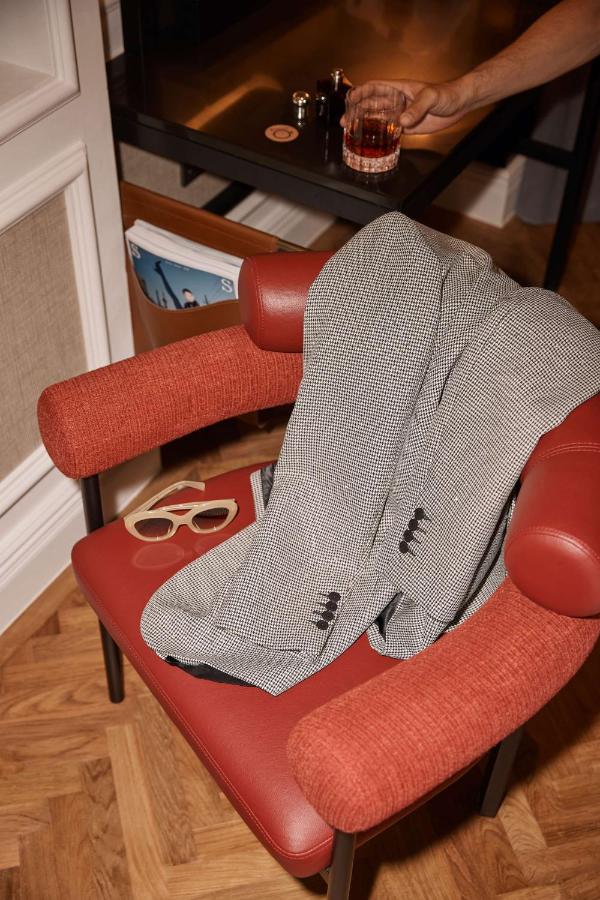 13 Most Unique Hotels in Singapore for an Incredible Holiday 30 A gray houndstooth blazer draped over a red chair with beige sunglasses on the seat evokes the sophisticated style found in unique hotels in Singapore. In the background, a hand holds a glass of dark liquor by a black desk with a magazine underneath.