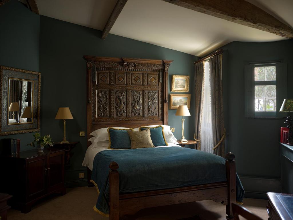 15 Best Boutique Hotels in London, UK 38 A cozy bedroom reminiscent of boutique hotels in London, featuring dark green walls, a wooden bed with an ornate carved headboard, gold-framed artwork, two bedside tables with lamps, and a window with sheer white curtains letting in natural light.