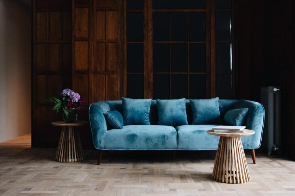 Best Boutique Hotels in San Sebastian 28 A modern living room, inspired by boutique hotels in San Sebastian, features a blue velvet sofa with four pillows, two round wooden side tables, a vase of flowers on the left table, and a book on the right against a wooden paneled wall.
