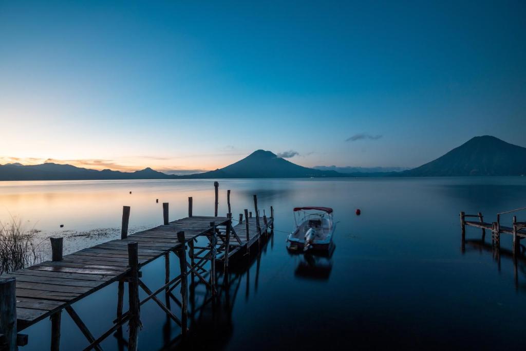 8 UNIQUE Places Where to Stay in Lake Atitlán 5 A wooden dock extends over a calm lake at dusk, with a small boat moored nearby. Mountains and a volcano silhouette the sky—an ideal scene for visitors seeking where to stay in Lake Atitlan.