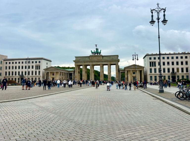 People walk and gather in front of the Brandenburg Gate in Berlin, Germany, on a cloudy day. The historic monument stands near modern buildings—just a short ride from the boutique hotels in Berlin Kreuzberg. Street lamps and bicycles are visible on the right.