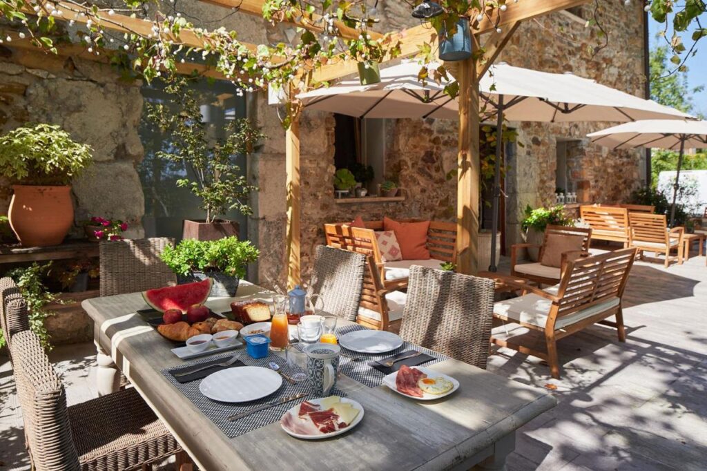 Best Boutique Hotels in San Sebastian 7 A sunny outdoor patio at one of the boutique hotels in San Sebastian features wooden furniture, a breakfast table with watermelon and ham, potted plants, and umbrellas providing shade next to a rustic stone building.