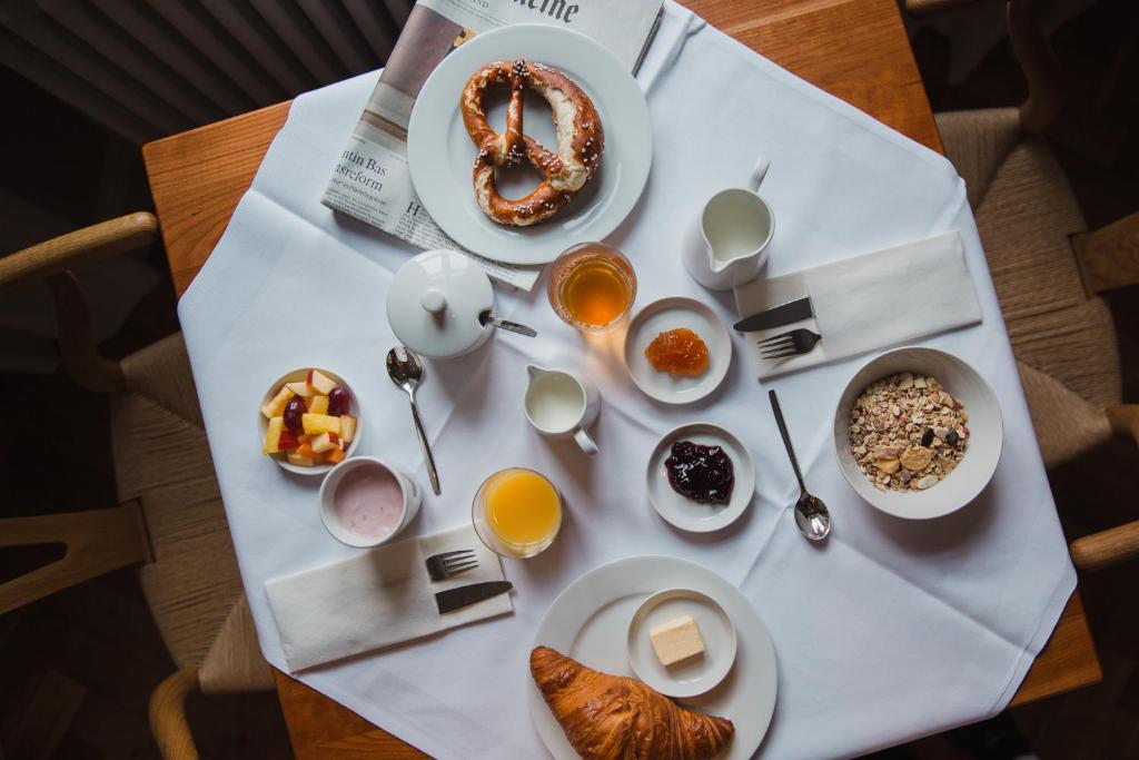 Authentic Boutique Hotels in Munich 26 A breakfast table at boutique hotels in Munich features a white tablecloth holding a croissant with butter, pretzel, cereals, fruit salad, yogurt, two glasses of juice, jams, tea, and newspapers—all arranged neatly with cutlery and napkins.