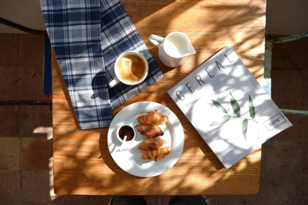 A wooden table with a blue checkered cloth, coffee, milk jug, mini croissants with jam, and a CEREAL magazine—an inviting scene reminiscent of mornings at boutique hotels in Costa Brava, sunlight dappled with leaf shadows.