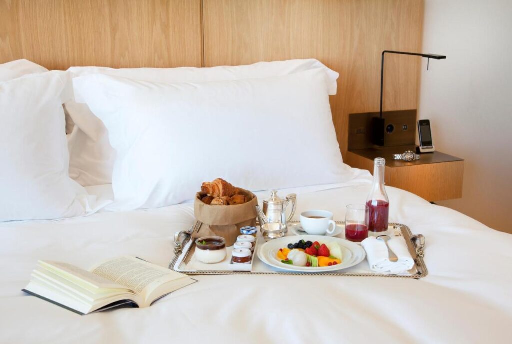 Best Boutique Hotels in San Sebastian 24 A breakfast tray on a neatly made bed with white linens, holding croissants, fruit, juice, coffee, and jam—just the kind of morning you might enjoy at boutique hotels in San Sebastian. An open book and a watch rest nearby on the bedside table.