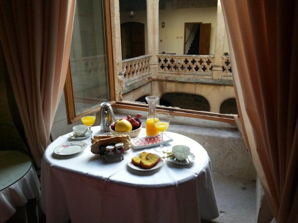 25 Weekend Trips from Madrid Max. 2h Away 28 A round table set for breakfast—with juice, coffee, bread, fruit, and pastries—sits by an open window overlooking a stone courtyard. The cozy, elegant scene evokes the charm of weekend trips from Madrid to historic towns.