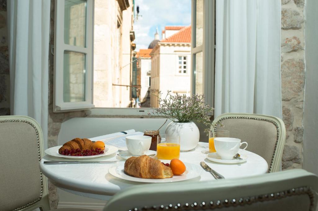 Most Unique Boutique Hotels in Dubrovnik 32 A small round table set for breakfast with croissants, fruit, juice, and coffee sits by an open window in one of the charming Boutique Hotels in Dubrovnik, letting in sunlight and offering a view of stone buildings with red-tiled roofs.