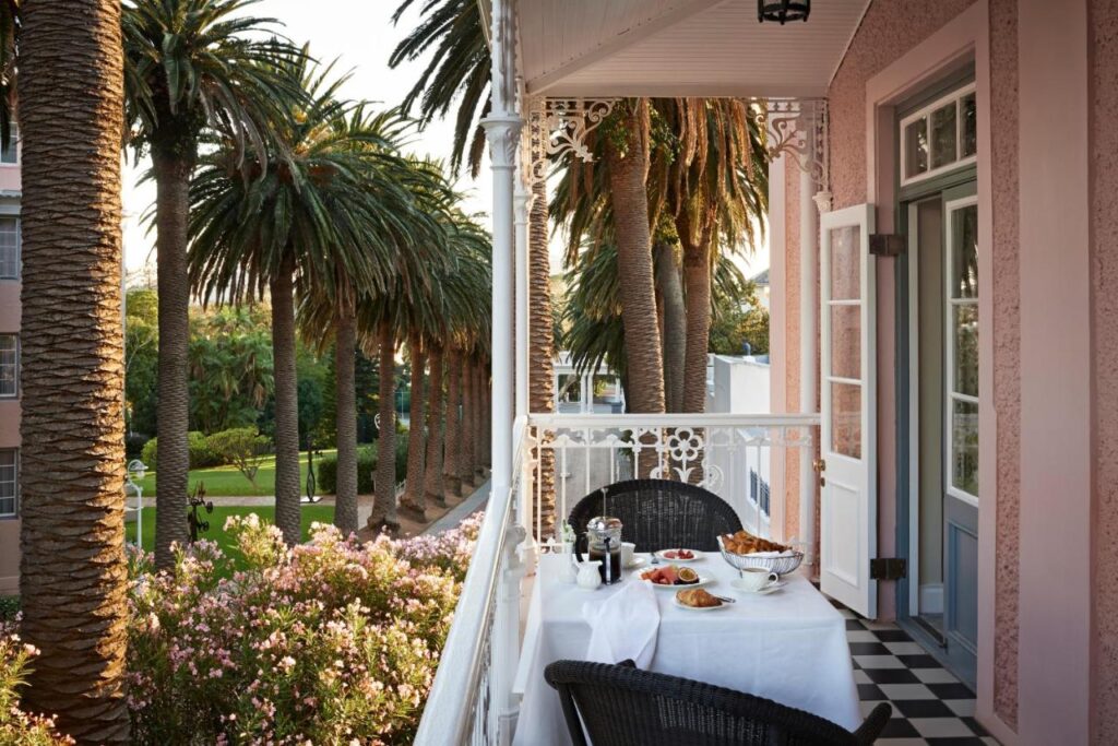 Unique Places to Stay in Cape Town 8 A balcony with black wicker chairs and a white table set for breakfast, overlooking a lush garden with palm trees. Sunlight filters through the trees—one of the truly unique places to stay in Cape Town, with pink walls and ornate white trim.