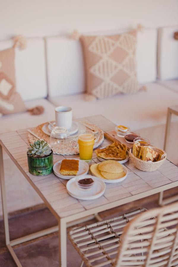 12 Charming Budget Riads in Marrakech 28 A small table in one of the budget riads in Marrakech is set for breakfast with juice, coffee, pastries, bread, spreads, a glass of water, and a potted plant. Cushioned seating with patterned pillows is in the background.