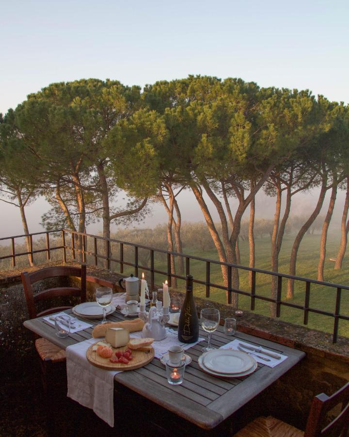 7 Best Agriturismos in Tuscany 5 A rustic outdoor dining table is set with plates, glasses, wine, bread, cheese, and candles—evoking the charm of the best agriturismos in Tuscany. Tall pine trees rise behind in a foggy sunrise or sunset, creating a peaceful atmosphere.