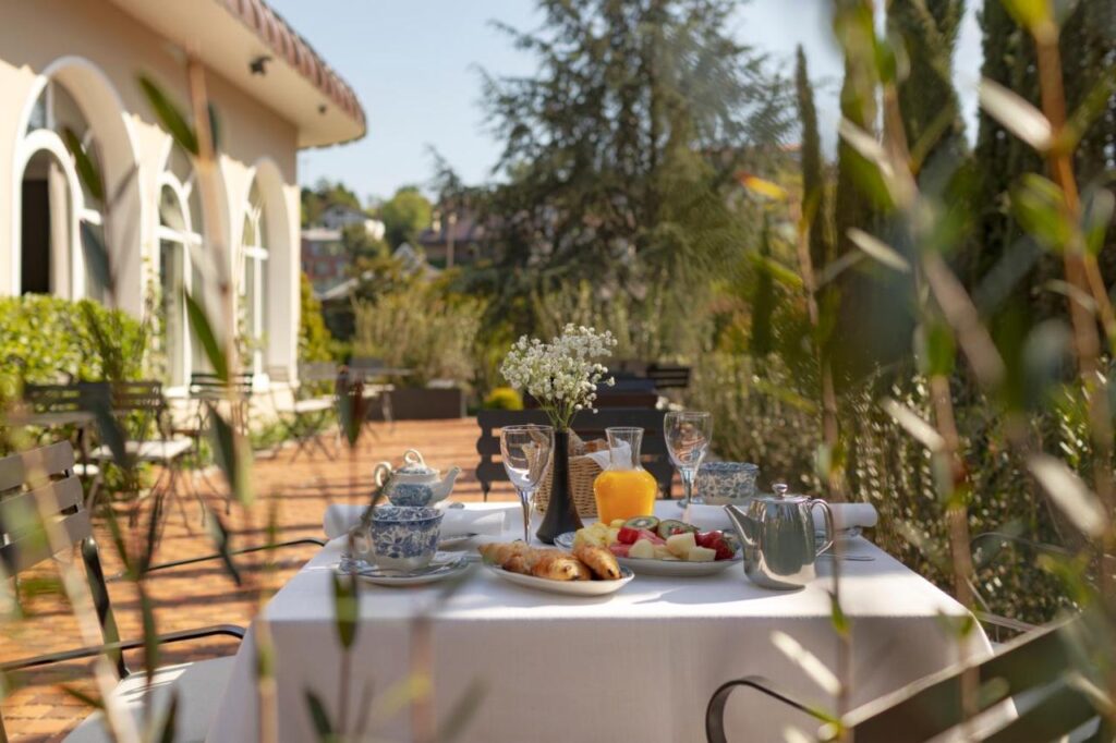 Best Boutique Hotels in San Sebastian 13 A table set for breakfast with tea, croissants, fruit, juice, and flowers sits on a sunny outdoor terrace surrounded by greenery—evoking the charming ambiance of boutique hotels in San Sebastian—next to a building with arched windows.
