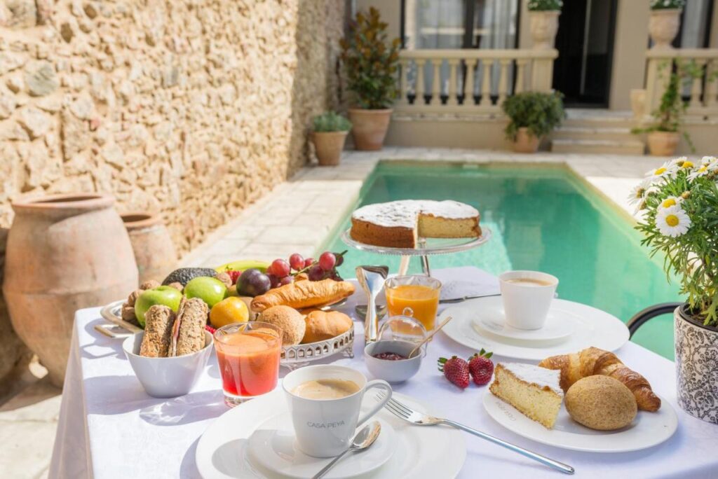 A table set for breakfast by a pool at one of the boutique hotels in Costa Brava features coffee, juice, pastries, fruit, cake, strawberries, and bread, with sunlight illuminating the cozy outdoor setting and flowers decorating the table.
