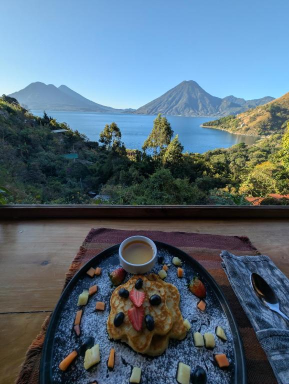 8 UNIQUE Places Where to Stay in Lake Atitlán 24 A breakfast plate with pancakes, fruit, and syrup sits on a wooden table. In the background, discover a scenic view of Lake Atitlan’s green hills and mountains—a perfect start for those seeking where to stay in Lake Atitlan.
