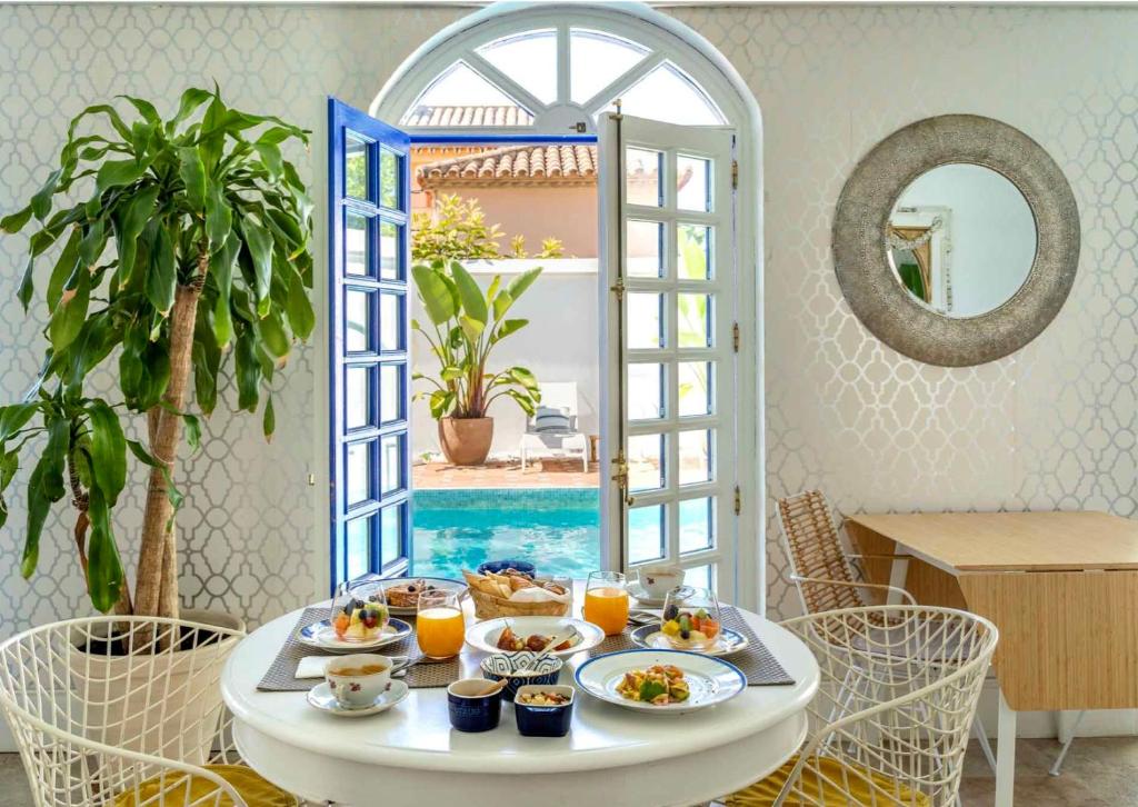 Adults-Only Hotels in Costa del Sol 19 A bright, airy dining room with a round table set for breakfast, reminiscent of exclusive adult only hotels in Costa del Sol. French doors open to a pool and leafy patio, while modern decor and a large potted plant complete the cozy scene.