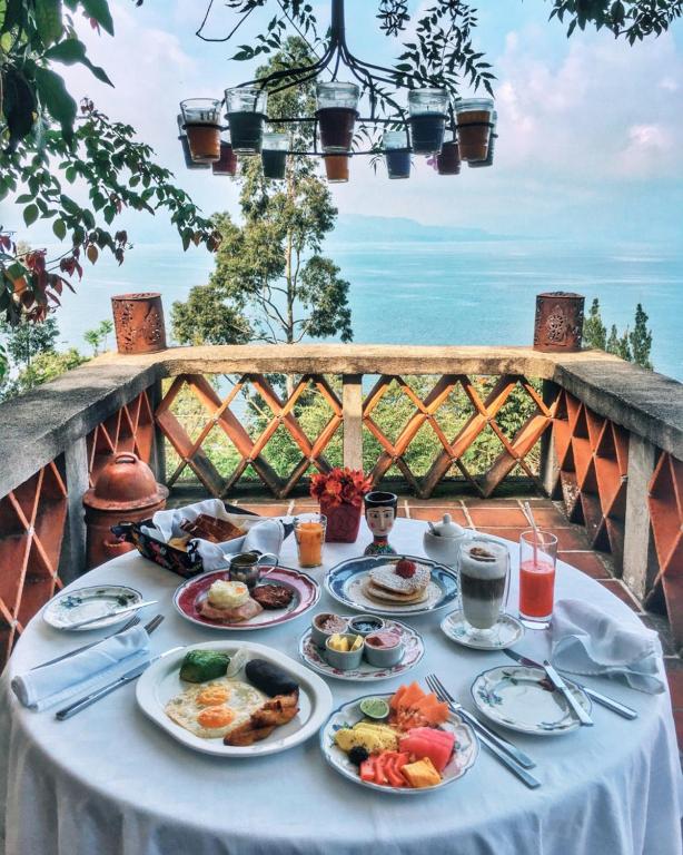 8 UNIQUE Places Where to Stay in Lake Atitlán 14 A round table is set with a variety of breakfast dishes, drinks, and fruit, overlooking a scenic ocean view from a terrace surrounded by greenery—a setting reminiscent of the best spots for where to stay in Lake Atitlan.