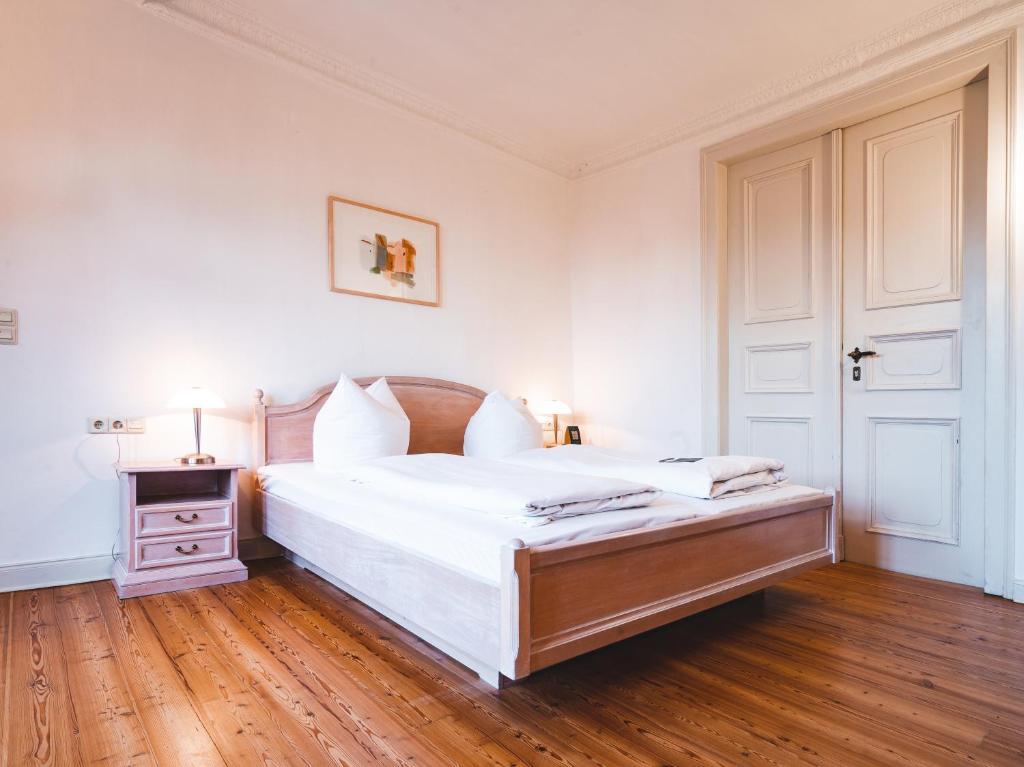 FAIRYTALE Castle Hotels in Bavaria 18 Bright bedroom with wooden floor, a double bed with white linens, two bedside tables with lamps, and framed artwork; large double doors are visible on the right, evoking the elegance found in castle hotels in Bavaria.