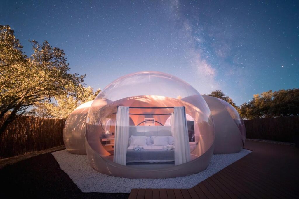 25 Weekend Trips from Madrid Max. 2h Away 19 A transparent dome-shaped tent with a bed inside sits under a starry night sky, surrounded by trees and a wooden deck—perfect for cozy glamping and unforgettable weekend trips from Madrid.