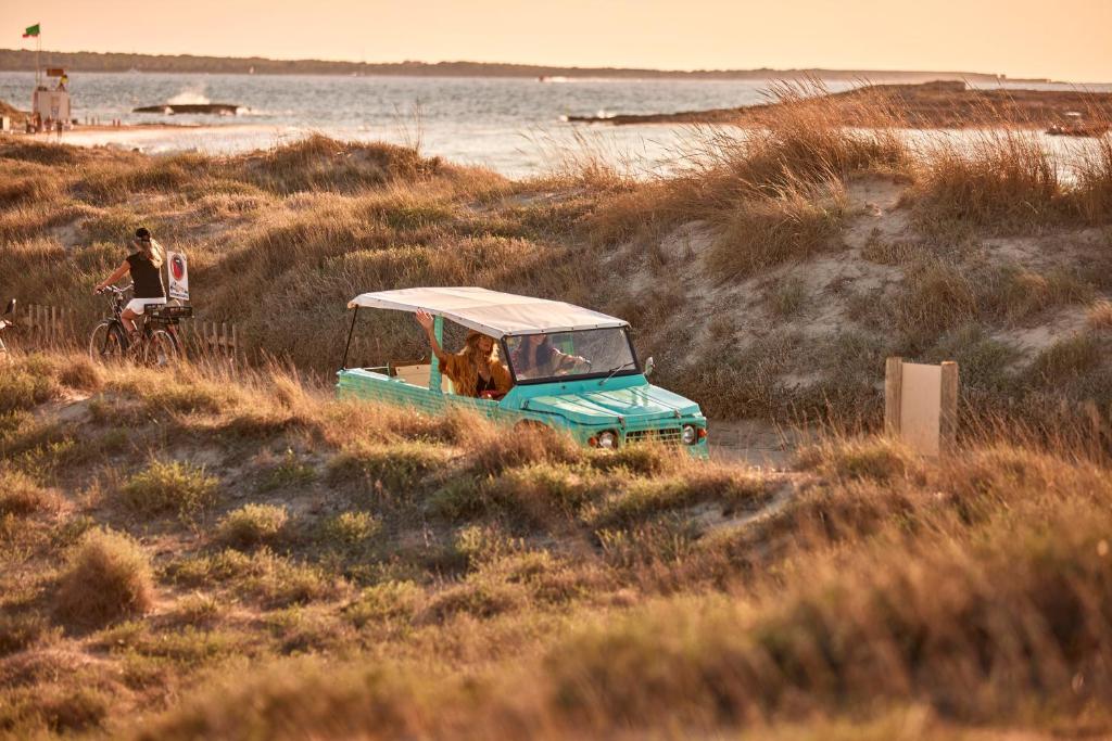 A turquoise open-top car cruises through sandy, grassy dunes near the beach at sunset, just steps from boutique hotels in Formentera. The sea shimmers in the background as a cyclist enjoys the golden sky.