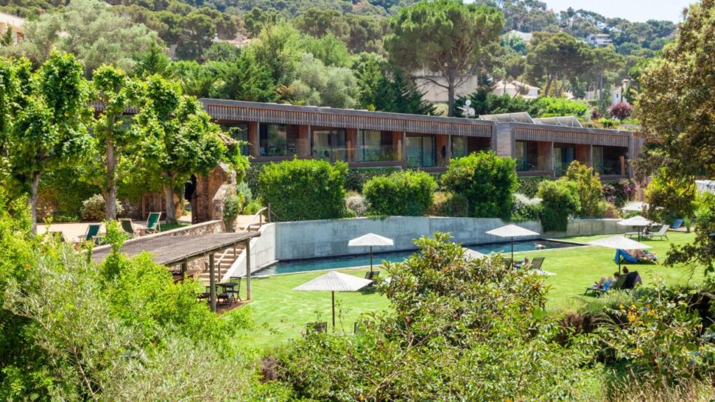 A modern, low-rise hotel surrounded by lush greenery features a long outdoor pool with sun loungers and umbrellas on a manicured lawn—an ideal escape for those seeking boutique hotels in Costa Brava. Trees and shrubs add privacy and shade.