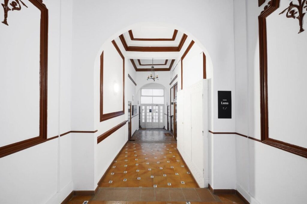 Where to Stay in Madrid: 14 Best Hotels 31 A hallway with white walls and brown trim, a brown-tiled floor, and a chandelier overhead leads to glass double doors. A black sign reads Luna 1.7—an inviting spot for those seeking where to stay in Madrid first time.